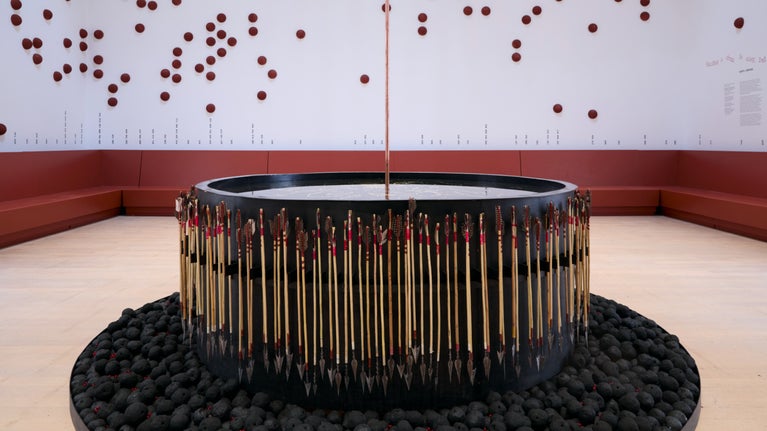 A sculpture with what looks like a large, black drum surrounded by spears pointing downward. At the base of the drum there is a cylinder of pieces of coal. The work sits inside an otherwise empty room.